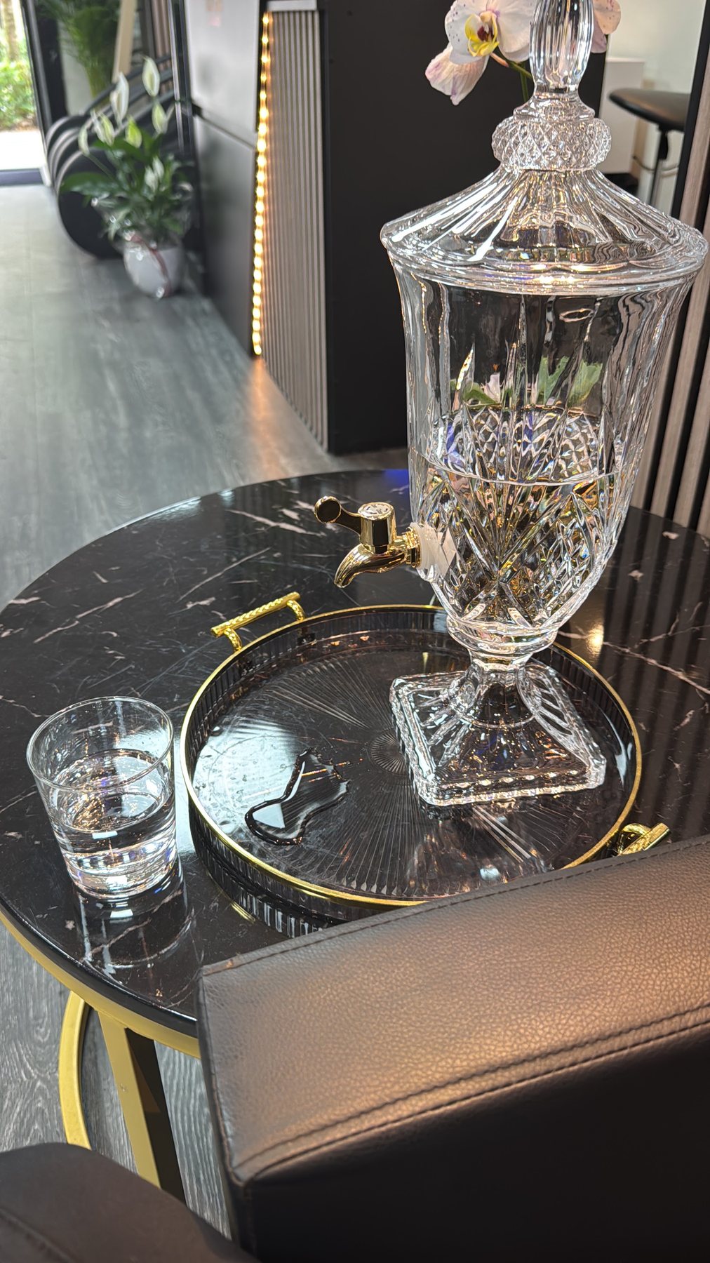 Een glazen drankdispenser en een glas water op een zwart marmeren tafel met goud